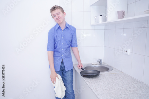 A cute teenager boy washing dishes at white kitchen alone with sad bad unhappy face