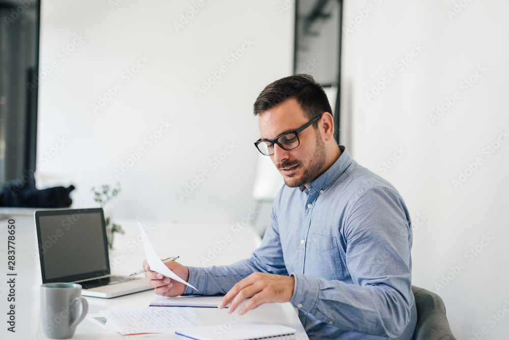 Happy young businessman looking at papers graphs and charts using laptop at his office desk Happy young businessman looking at papers graphs and charts using laptop at his office desk