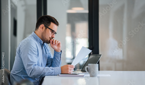 Serious pensive thoughtful young businessman or entrepreneur in modern contemporary office looking at and working with laptop and paper photo