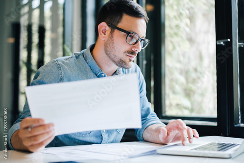 Serious pensive thoughtful focused young casual business accountant bookkeeper in office looking at and working with laptop and income tax return papers and documents photo