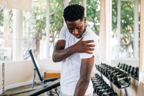 Side view of muscular African American man standing and suffering from shoulder pain during workout with dumbbells. Sportsman holding sore shoulder in gym photo