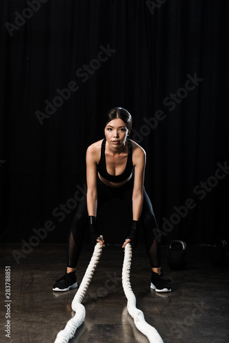 selective focus of beautiful sportswoman exercising with battle ropes on black