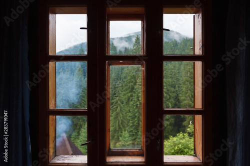 Wooden window behind which a mountain forest is visible. © Nataliia Hrihel