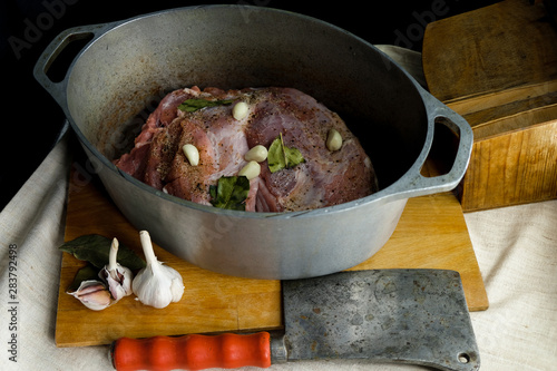 Piece of raw meat with condiments in a cauldron. Near the cauldron there is a chopper and garlic.