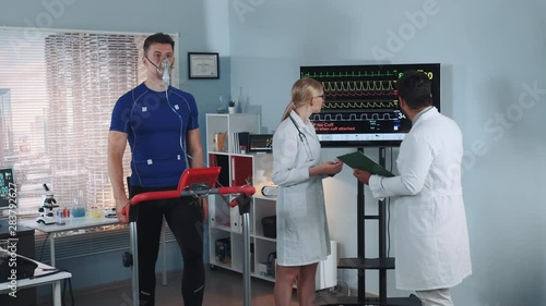Multiracial scientists examining the athlete's medical records while he stress testing in lab with skyscrapers in the background. In Scientific Sports Laboratory.