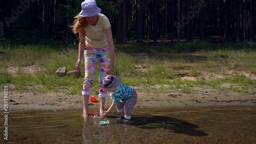 Mom with a small son on a sunny summer day launch a toy boat on the shore of a forest lake. Happy moments of motherhood and childhood