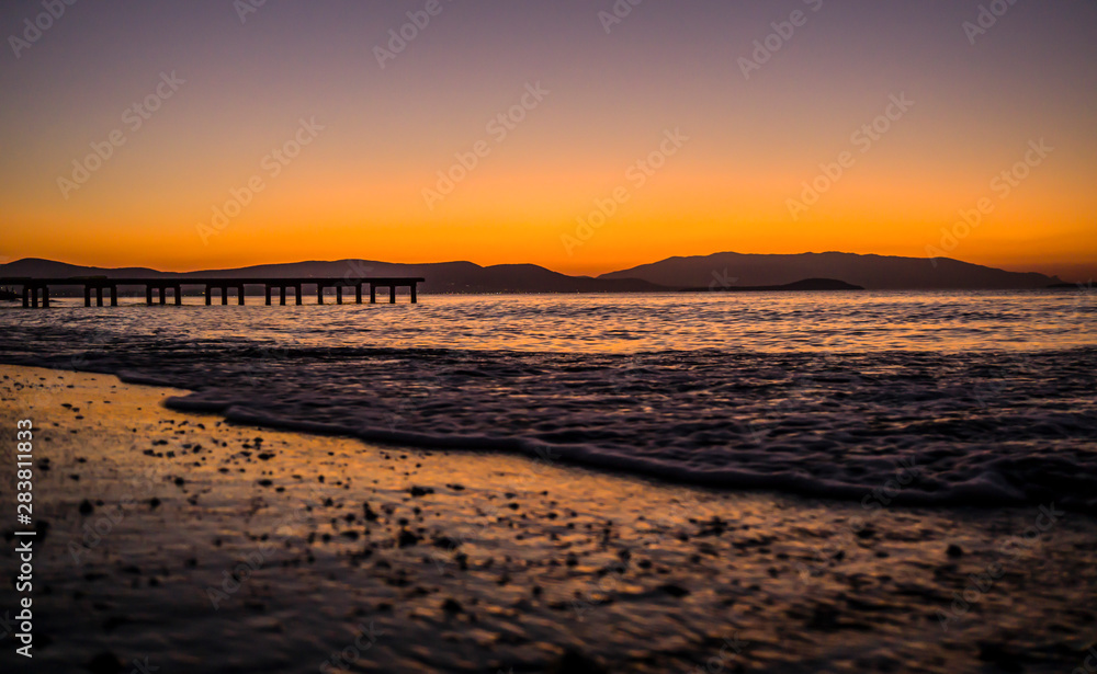 Dramatic sunset landscape at Urla, Izmir, Turkey. Beautiful blazing sunset over bright blue sea, pier, orange & purple sky above it with awesome golden rays of sun light reflection on calm waves.