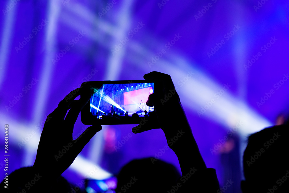 Unrecognizable people shoot photo and video on the mobile phone in front of the stage on the show. Dark blurred silhouettes of a concert crowd with their hands up in front of bright lights. Unrecognizable people shoot photo and video on the mobile phone in front of the stage on the show. Dark blurred silhouettes of a concert crowd with their hands up in front of bright lights.