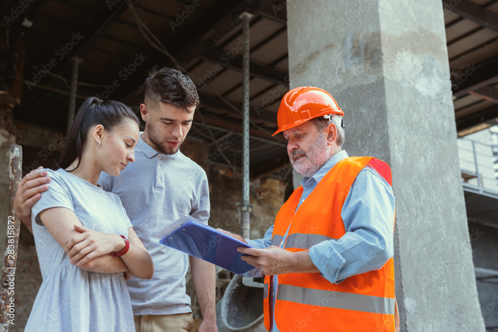 Foreman or achitect engineer shows future house, office or store design plans to a young couple. Meeting at the construction site to talk about facade appearance, interior decoration, home layout.