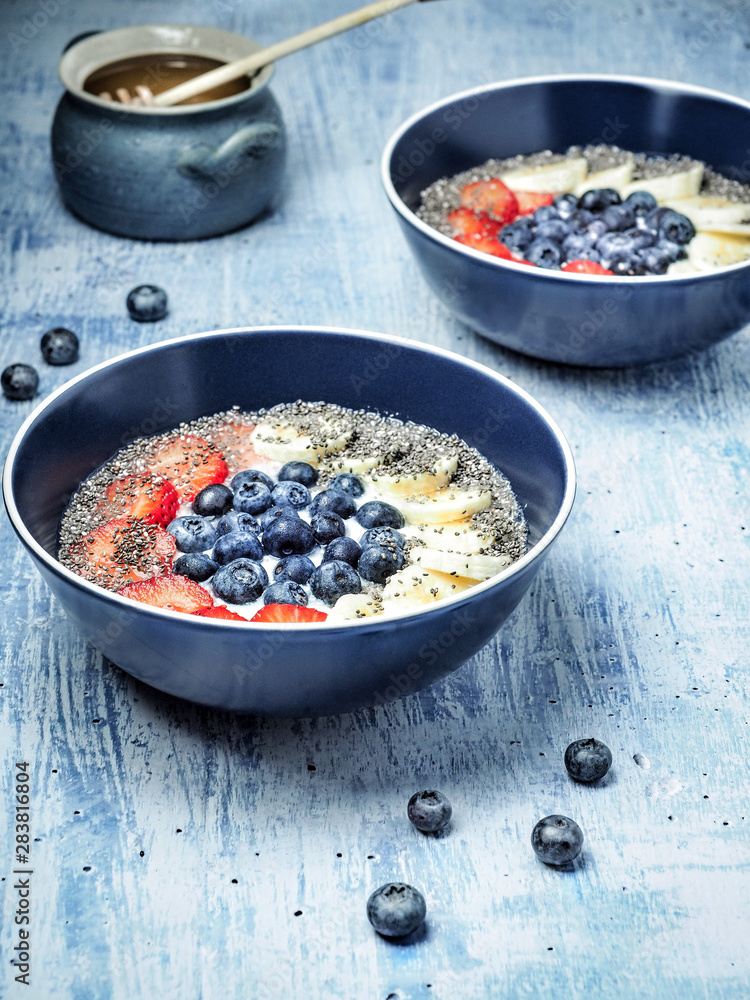 Two bowls of yoghurt and fruits - strawberries, blueberries and banana, cup of honey on blue table. Overhead shot. Two bowls of yoghurt and fruits - strawberries, blueberries and banana, cup of honey on blue table. Overhead shot.