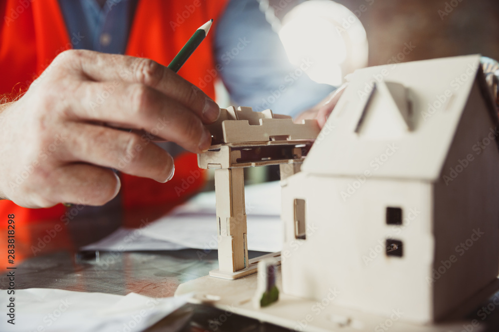 Close up photoshot of male architect-engineer making a model of future house for young family. Man working in the office with miniature, drawings, blueprint. First home, industrial, building concept. Close up photoshot of male architect-engineer making a model of future house for young family. Man working in the office with miniature, drawings, blueprint. First home, industrial, building concept.