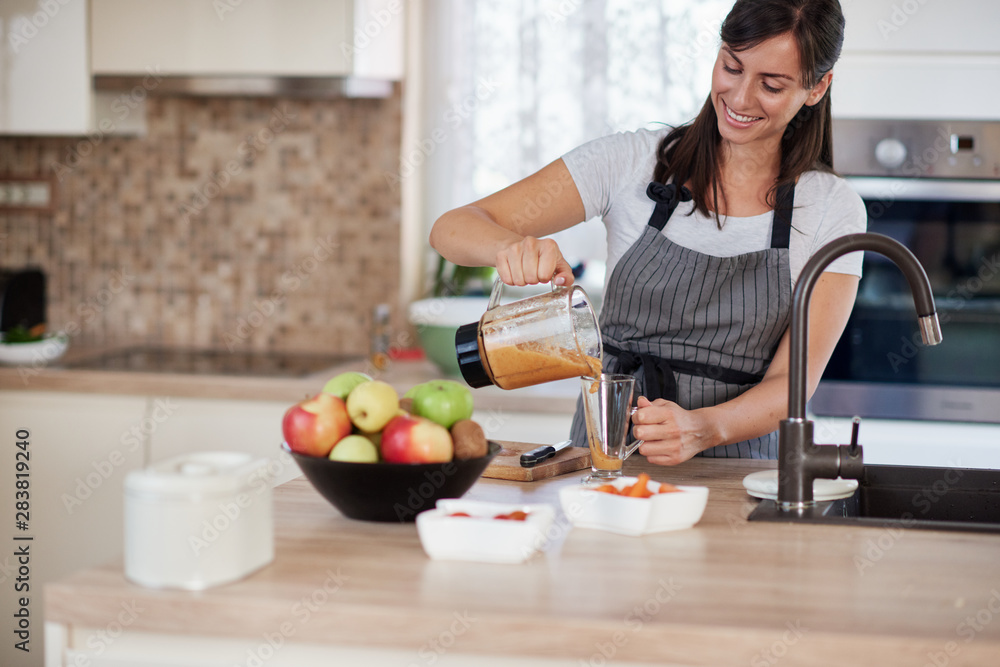 Beautiful smiling Caucasian woman in apron standing in kitchen and pouring fresh smoothie into glass.