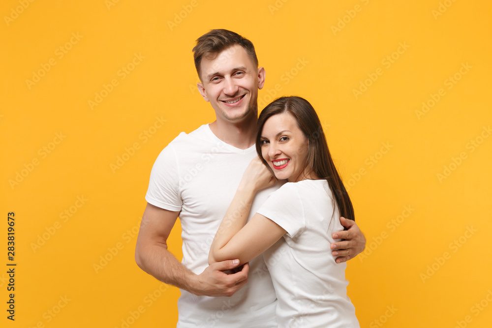 Joyful young couple two friends guy girl in white empty blank design t-shirts posing isolated on yellow orange wall background studio portrait. People lifestyle concept. Mock up copy space. Hugging. Joyful young couple two friends guy girl in white empty blank design t-shirts posing isolated on yellow orange wall background studio portrait. People lifestyle concept. Mock up copy space. Hugging.