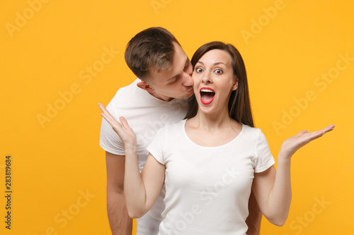 Excited young couple two friends guy girl in white empty blank design t-shirts posing isolated on yellow orange wall background. People lifestyle concept. Mock up copy space. Kissing, spreading hands.