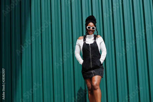 African woman in denim overalls skirt, white sunglasses posed against green steel wall.