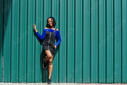 African woman in denim overalls skirt, eyeglasses posed against green steel wall.