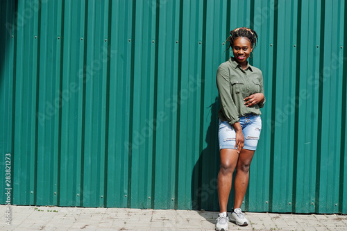 African woman with dreads hair, in jeans shorts posed against green steel wall.