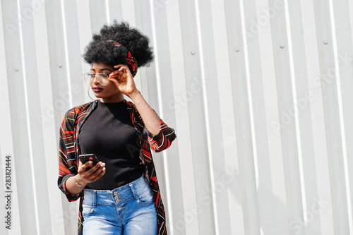African woman with afro hair, in jeans shorts and eyeglasses posed against white steel wall with mobile phone in hand.