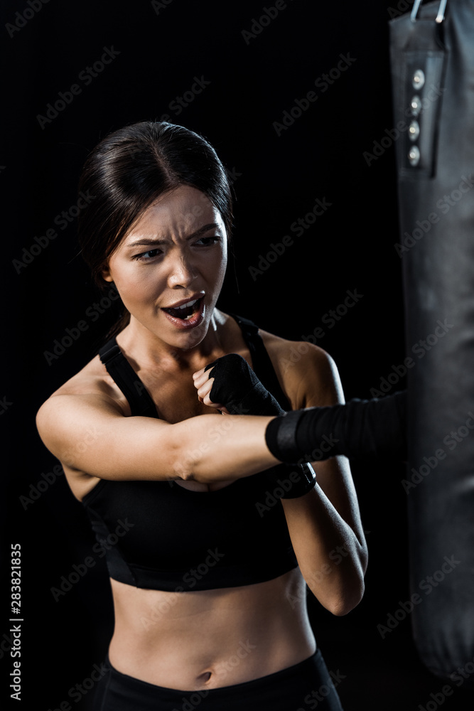selective focus of emotional sportswoman boxing near punching bag isolated on black