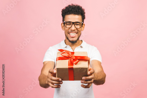 Cheerful smiling african american millennial guy in eyewear holding wrapped present box. Happy black young man congratulating, giving birthday gift, isolated on pink background. © denis_vermenko