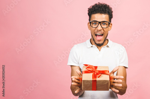 Cheerful smiling african american millennial guy in eyewear holding wrapped present box. Happy black young man congratulating, giving birthday gift, isolated on pink background. © denis_vermenko