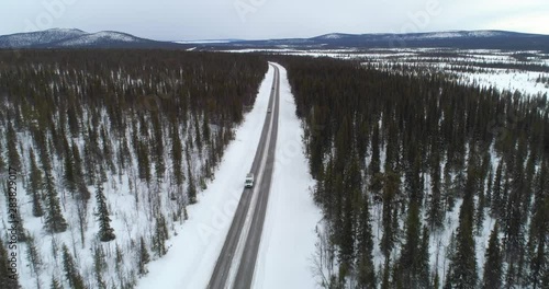 Car on lappland road, C4k aerial, reverse, drone shot, following a van, driving between spruce forest, in arctic wilderness, on a cloudy, winter day, near Kiruna, in Norrbotten, Sweden photo
