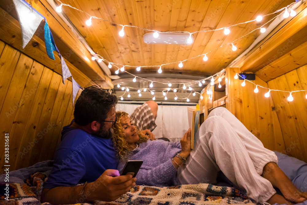 Travel and romantic night inside a wooden restored beautiful van for happy adult caucasian couple in love relaxing with phone and paper book - wanderlust and traveler people together