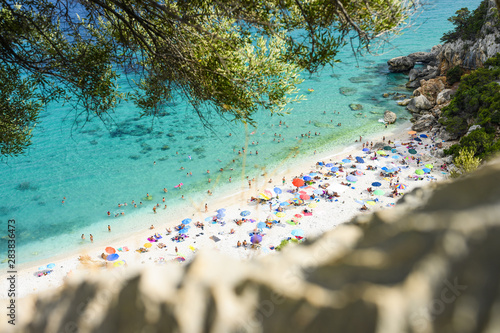 Fototapeta Naklejka Na Ścianę i Meble -  (Selective focus) View from above, stunning view of a beautiful beach full of beach umbrellas and people sunbathing and swimming on a turquoise water. Cala Gonone, Sardinia, Italy.