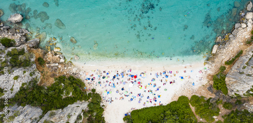 View from above, stunning aerial view of a beautiful beach full of beach umbrellas and people sunbathing and swimming on a turquoise water. Cala Gonone, Sardinia, Italy.