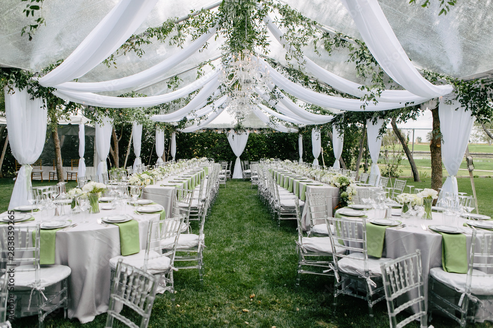 Outdoor summer wedding tent decorated with hanging fabric, greenery, and crystal chandeliers, wedding reception tables, green accent color - obrazy, fototapety, plakaty 