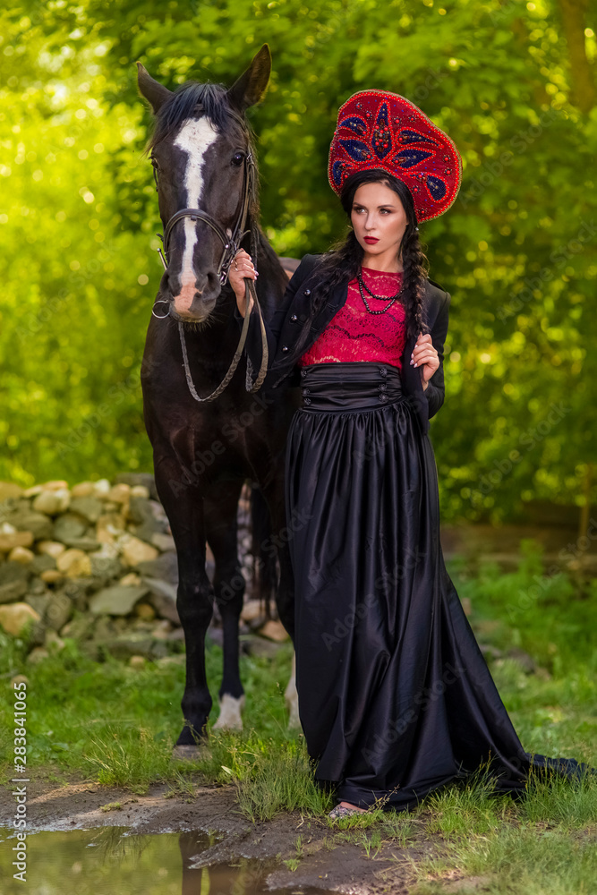 Model in Russian Kokoshnik Holding Thoroughbred Horse Against Nature Background. Model in Russian Kokoshnik Holding Thoroughbred Horse Against Nature Background.