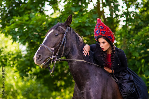 Gorgeous Model in Russian Style Kokoshnik Straddling On the Thoroughbred Horse. Posing Against Nature Background.