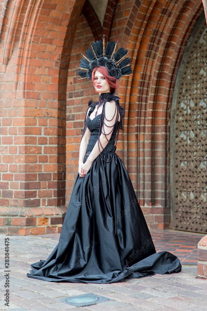 Portrait of Tranquil Gothic Girl in Long Black Dress. Wearing Artistic Feather Crown with Roses. Posing Against Old Castle Gates.