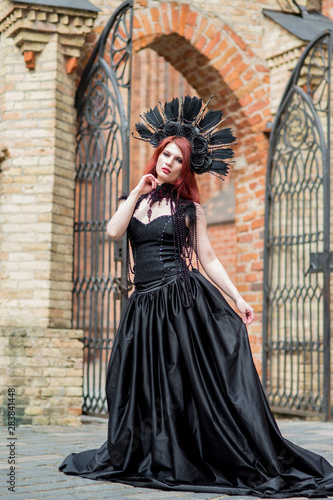 Portrait of Gothic Caucasian Woman in Black Dress and Artistic Feather Crown. posing Against Old Castle Gates