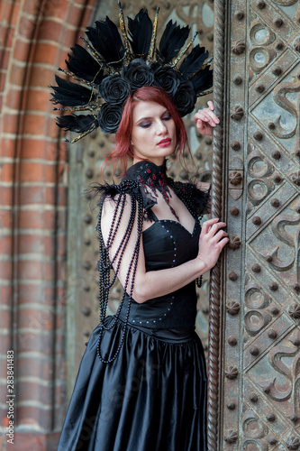 Gothic Girl in Long Black Dress. Wearing Artistic Feather Crown with Roses. Posing Against Old Castle Gates.