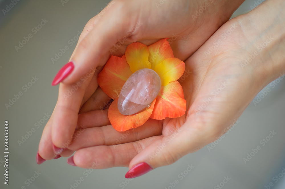 Female hand with red manicure holding transparent violet amethyst yoni egg for vumfit, imbuilding or meditation inside orange and yellow gladiolus flower.