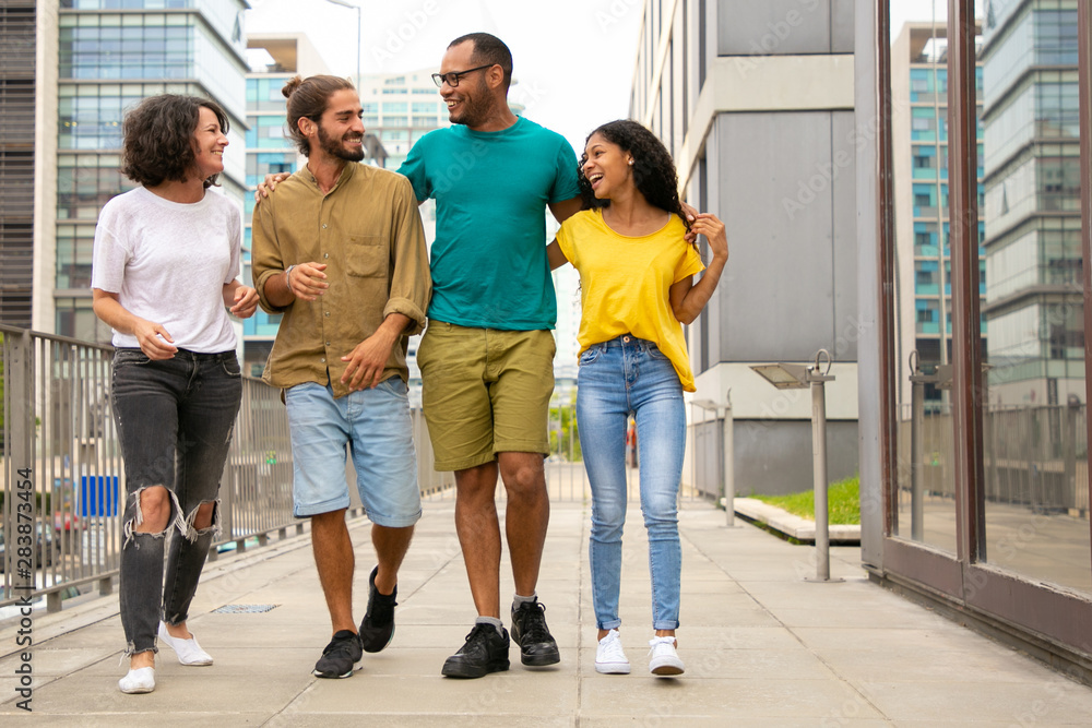Happy close friends enjoying outdoor walk. Young multiethnic people walking down urban street, hugging each other, talking and laughing. Hangout concept