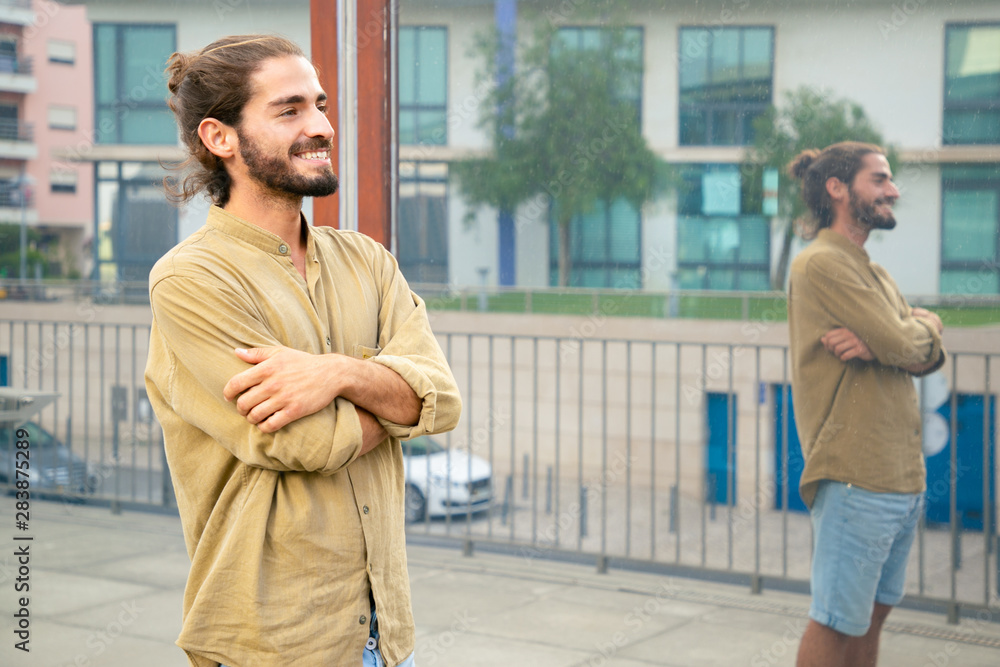 Happy hipster guy waiting someone outside. Young man in casual with bun and stubble standing in city street, looking into distance and smiling. Waiting outdoors concept