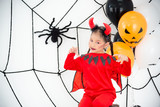 Pretty asian girl in red devil costume with horn sitting in room decorated for Halloween festival.