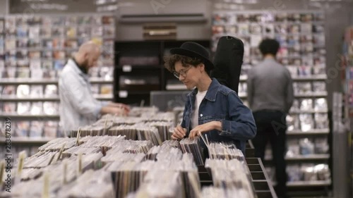 Panning medium shot of curly Caucasian female musician, wearing bowler hat, denim jacket and glasses, with guitar gig bag on shoulder, browsing through selection of vinyls in record store