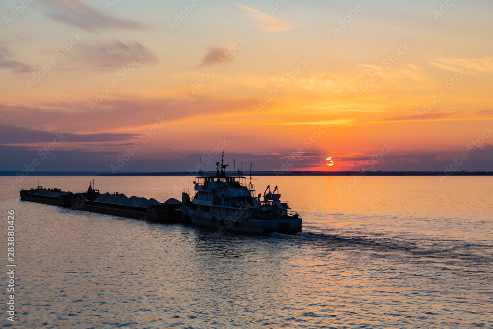A tug on the river pushes a barge with sand or gravel. Sunset on the Volga river in Russia in July. Orange sun in the sky. Picturesque nature. Summer evening. River cruise A tug on the river pushes a barge with sand or gravel. Sunset on the Volga river in Russia in July. Orange sun in the sky. Picturesque nature. Summer evening. River cruise