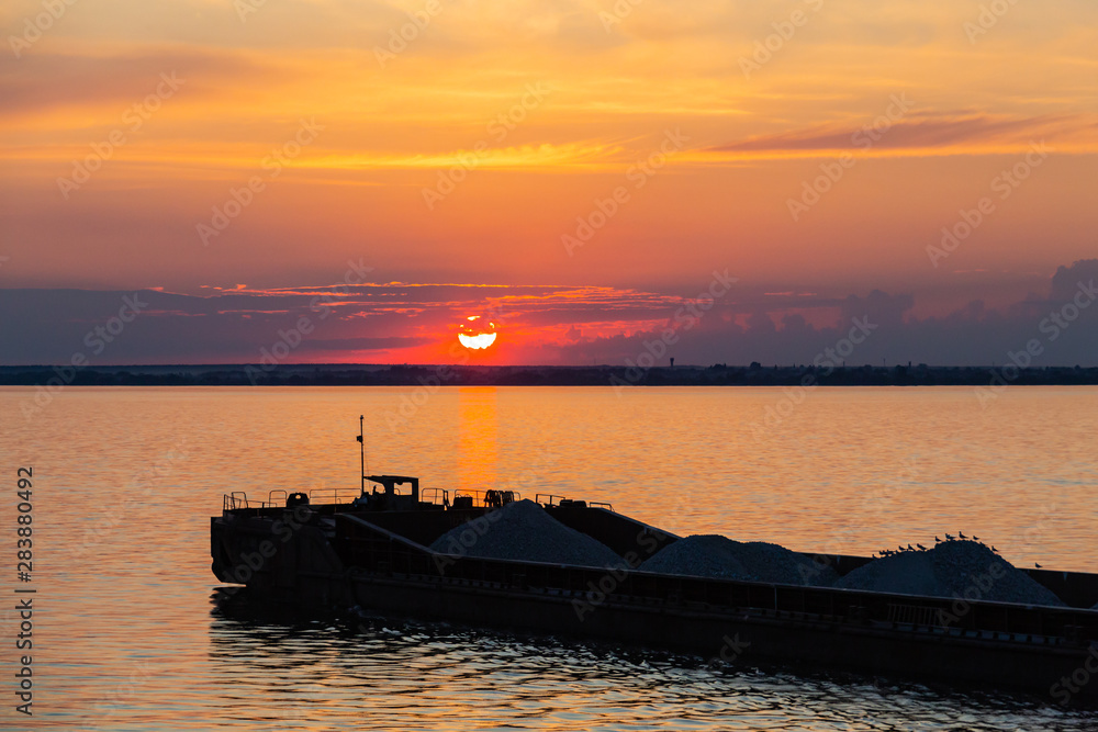 A tug on the river pushes a barge with sand or gravel. Sunset on the Volga river in Russia in July. Orange sun in the sky. Picturesque nature. Summer evening. River cruise