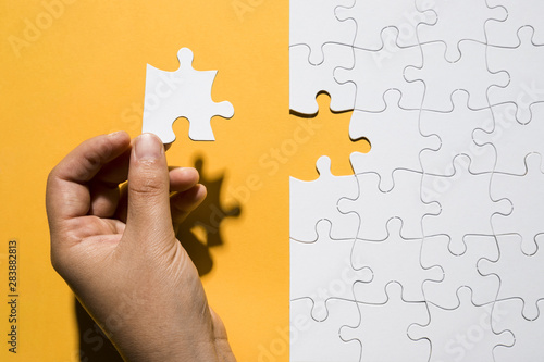 Human hand holding puzzle piece over white puzzle grid over yellow backdrop