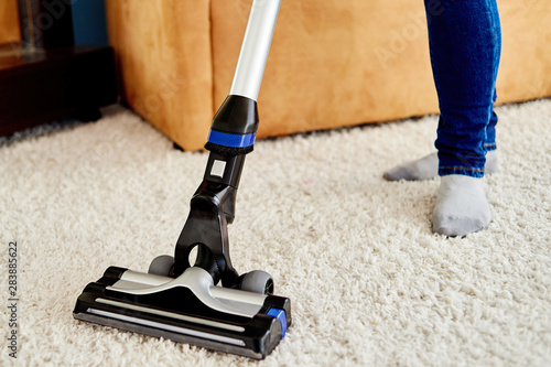 Close up of young woman in jeans cleaning carpet with vacuum cleaner in living room, copy space. Housework, household, spring-cleaning and chores concept. House cleaning