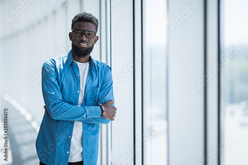 Thoughtful young african businessman with arms crossed in office against panoramic window