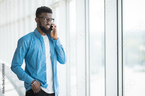 Handsome serious African American man in suit standing near window in office looking away at cityscape while speaking on mobile phone