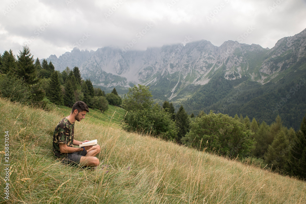 Caucasian man sitting in the grass and reading a book in a mountain meadow. Beautiful natural landscape behind. Human and nature connection concept