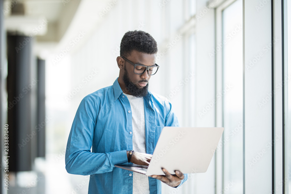 Love my job. Waist-up portrait of positive attractive african man in glasses is standing in office and holding modern laptop near panoramic windows Love my job. Waist-up portrait of positive attractive african man in glasses is standing in office and holding modern laptop near panoramic windows