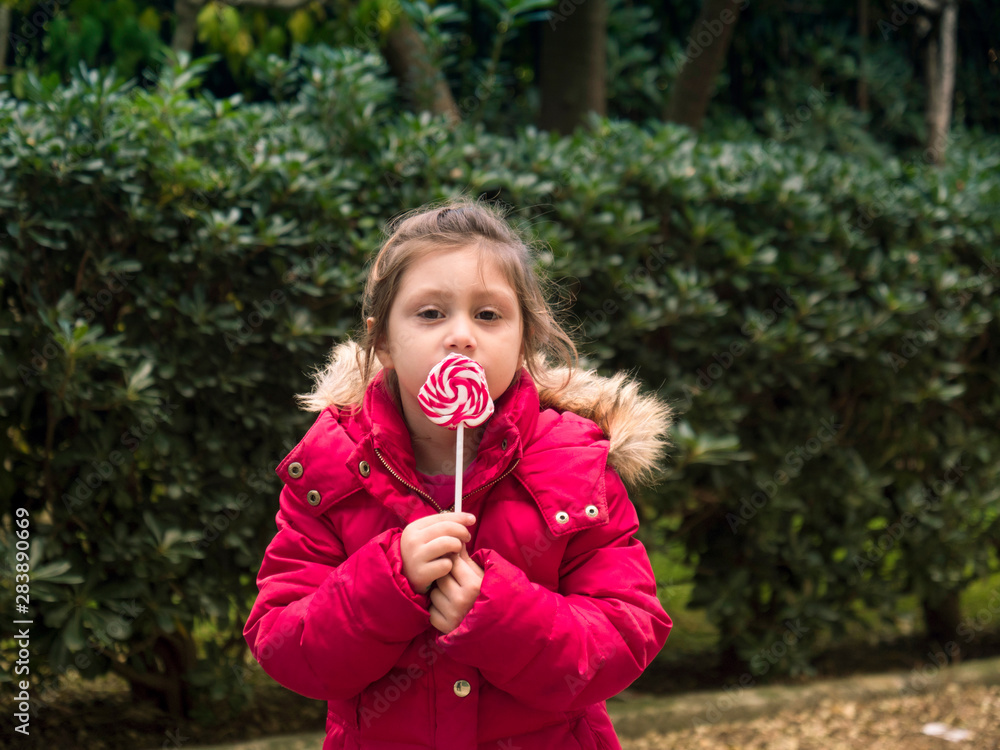 Happy young little child girl kid bite sweet lollipop candy. Portrait of a smiling girl with a lollipop in summer time Happy young little child girl kid bite sweet lollipop candy. Portrait of a smiling girl with a lollipop in summer time