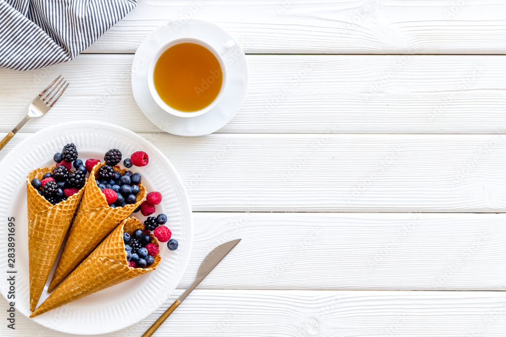 Light breakfast with fresh berries in waffle cones and cup of tea served white wooden table background top view mockup